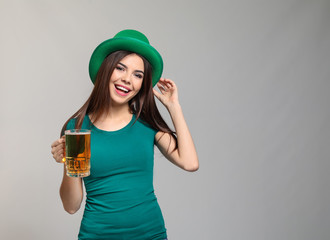 Beautiful young woman in green hat and with mug of beer on grey background. St. Patrick's Day...