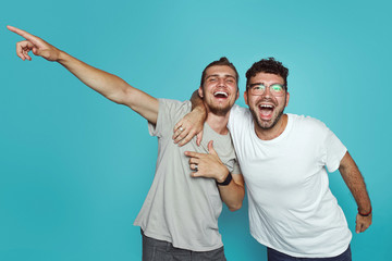 Image of two happy hipsters guys pointing fingers away isolated over light blue background. Friendship concept.