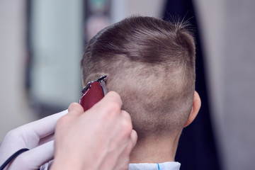Barber is using clipper while making hair tattoo for a cute European boy in barbershop.