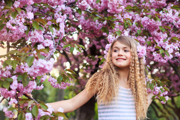 Pretty little girl with curly hair in pink dress walking in blossom cherry garden in sunny spring day