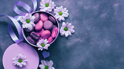 Macaroons of purple and pink color in a gift box decorated with flowers on a blue background. Overhead view, copy space. International Women's Day March 8th.