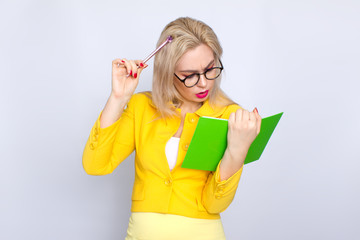 Close-up portrait of amazing blonde woman holding notebook, book, literature, in hands arms and pen pencil in yellow suit over white background in studio