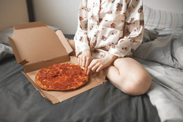 Girl sits in bed in the bedroom of the house, takes a piece of pizza from the box. Woman in pajamas eats on the bed with an appetizing pizza from the delivery. Fast Food, Pizza Concept.