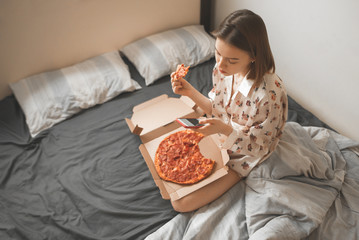 Attractive girl in a pajama sits on a bed with a pizza box on her lap, uses a smartphone and eats a...