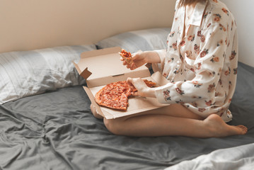 Woman in a pajama sits on the bed, holds a pizza box on her knees, and a piece in her hands. Girl sits on a dark sheet and eats fast food. Copyspace