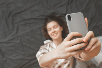 Pretty girl dressed in pajama lays on bed and makes a sephi on a smartphone. Top view, focus on smartphone. Copyspace