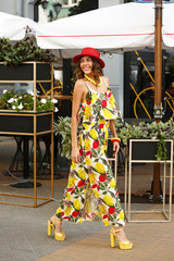 Bright stylish girl dressed in a colorful dress, red hat and yellow high heels is walking in the...