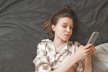 Attractive girl in pajamas, lying on the bed, holding a smartphone in her hands and looking into the camera. Beautiful girl with phone in hand, resting on bed and looking into camera. Copyspace