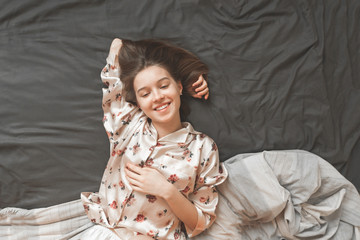 Happy girl in a pajama lays on the bed in the bedroom and smiles frankly. Joyful girl resting at home in bed, top view.