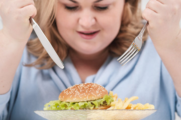 Young curvy fat woman in casual blue clothes on a white background at the table eagerly eating fast...