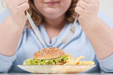 Young curvy fat woman in casual blue clothes on a white background at the table eagerly eating fast food, hamburger and french fries. Diet and proper nutrition.