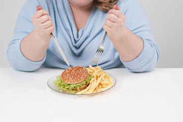 Young curvy fat woman in casual blue clothes on a white background at the table eagerly eating fast food, hamburger and french fries. Diet and proper nutrition.