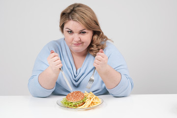Young curvy fat woman in casual blue clothes on a white background at the table eagerly eating fast...