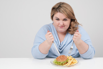 Young curvy fat woman in casual blue clothes on a white background at the table eagerly eating fast food, hamburger and french fries. Diet and proper nutrition.