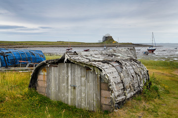 Weathered hull of shed made of overturned boat cut in half on Holy Island with Lindisfarner Castle ruins under renovation England UK