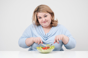 Young lush fat woman in casual blue clothes on a white background at the table and eats a vegetable salad with tomatoes. Diet and proper nutrition.