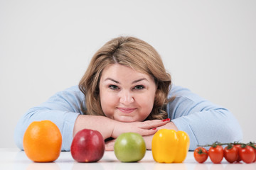 Fototapeta na wymiar Young lush fat woman in casual blue clothes on a white background at the table, in front of her in a row laid out vegetables and fruits. Diet and proper nutrition.