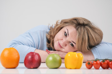 Fototapeta na wymiar Young lush fat woman in casual blue clothes on a white background at the table, in front of her in a row laid out vegetables and fruits. Diet and proper nutrition.