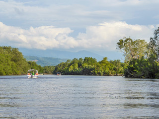 A boat tour through mangrove forest in Kota Kinabalu, Saba, Malaysia. Two boats in front. Green trees to the sides, river in the middle. High mountains in the back. Disturbed surface of the river.