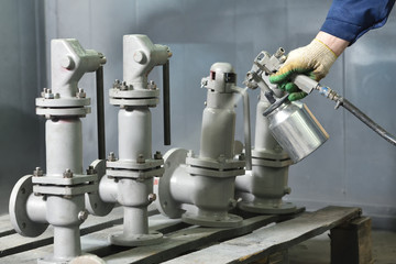 A worker at a factory in a special room paints parts of the valves from the spray gun. Paint valves in gray color, hand and spray gun close up