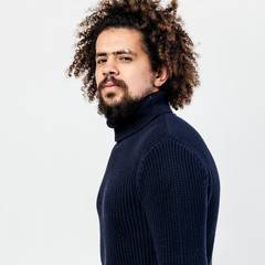 Dark-haired curly guy with a beard dressed in a black knitted turtleneck poses in the studio on the white background.