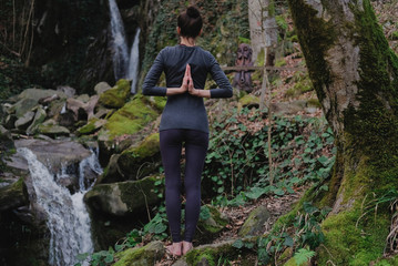 Young slim woman practicing yoga outdoors in moss forest on background of waterfall. Unity with nature concept. Girl standing with prayer hands