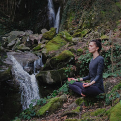 Young slim woman practicing yoga outdoors in moss forest on background of waterfall. Unity with nature concept. Girl meditates sitting