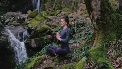 Naklejka na ściany i meble Young slim woman practicing yoga outdoors in moss forest on background of waterfall. Unity with nature concept. Girl meditates sitting