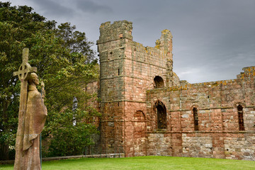 Life size statue of St Aidan first bishop of Lindisfarne facing ruins of the medieval priory on...