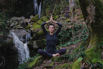 Young slim woman practicing yoga outdoors in moss forest on background of waterfall. Unity with nature concept. Girl meditates sitting