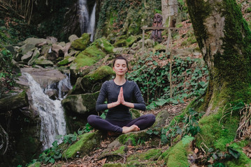 Young slim woman practicing yoga outdoors in moss forest on background of waterfall. Unity with...