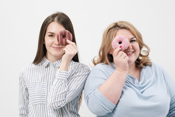 Two girls, thin and fat on a white background, are holding pink glazed donuts in their hands.