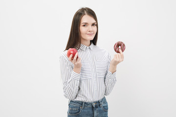 A slender beautiful brunette girl on a white background holding a pink glazed donut and a red apple.