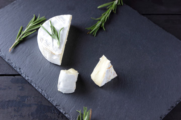 Camembert and rosemary on black stone board. Soft cheese with white mold on black background. Sliced cheese and rosemary branches on slate board