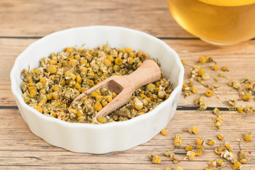 Dried chamomile in a white bowl and a glass of tea on wooden background.