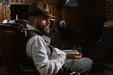 A man in hat wearing vintage suit holding pipe and glass of whiskey sitting on a big chair