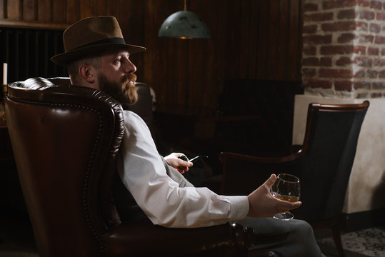 A man in hat wearing vintage suit holding pipe and glass of whiskey sitting on a big chair