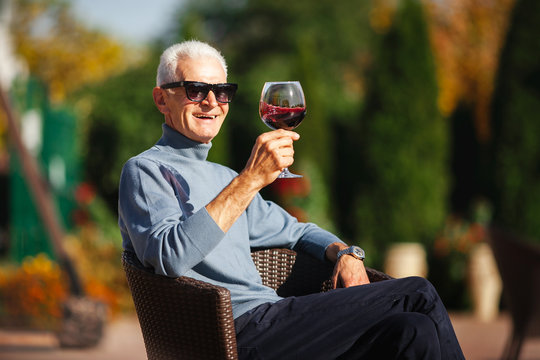 Relaxed old grey-haired man sit at the garden and enjoy glass of wine