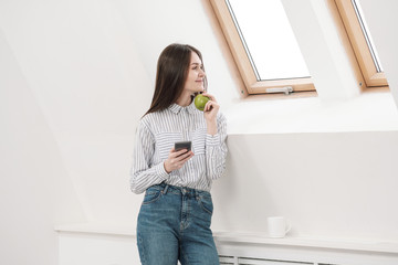 Slender brunette girl with long hair on a white background near the office window. Uses a smartphone and chatting and relaxing during a lunch break. Useful snack green apple.