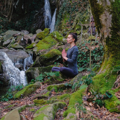 Young slim woman practicing yoga outdoors in moss forest on background of waterfall. Unity with nature concept. Girl meditates sitting