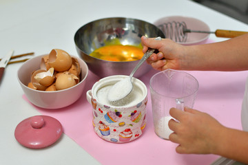 girl pours sugar into a measuring cup with a spoon. Little girl preparing cookies in kitchen at home. Cooking homemade food.