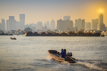 Lifestyle of villagers uses a small boat across the Chao Phraya river at Bang Krachao district pier with Bangkok city and row of barge boats background.