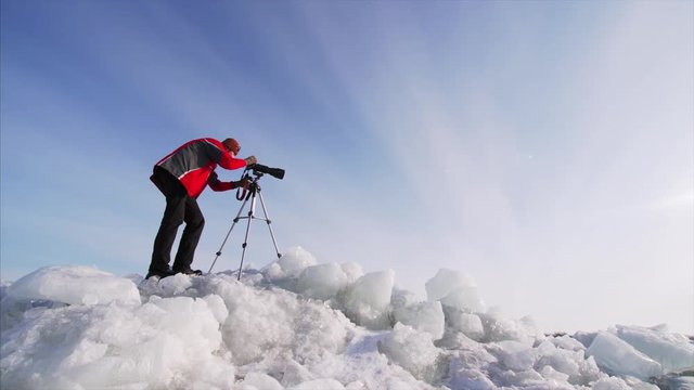 Sliding view from below of photographer with camera on ice blocks taking photos or video of frozen lake. Blue sky is on the background