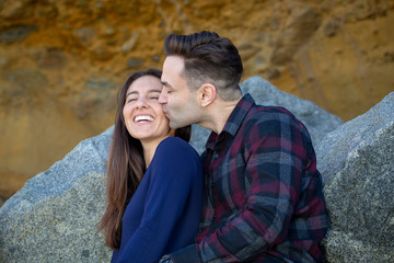Happy Millennial Couple Sitting on Rocks Boyfriend Kissing Girlfriend