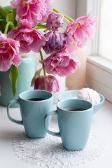Fototapeta na wymiar Two cups of black coffee and a delicate marshmallow for breakfast are next to a beautiful bouquet of pink tulips in a blue vase.