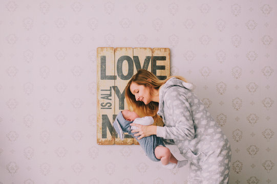 Indoor portrait of  hapy young girl in pijama holding, hugging her newborn baby boy near  wall with decorative plate. Mother take care of her infant child.  Motherhood concept. Love is all you need.