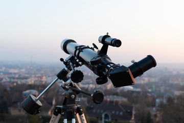 A professional refractor telescope mounted on a tripod, looking at a major city at sunset