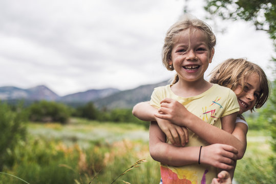 Portrait Of Happy Sisters Embracing While Standing On Landscape Against Cloudy Sky In Forest