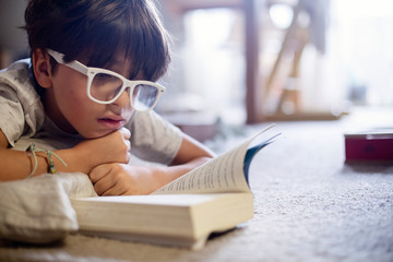 Boy wearing eyeglasses reading book while lying on carpet at home