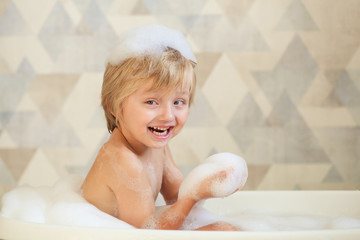 Little baby taking a bubble bath in a beautiful bathroom with a blue wall and flowers.  Shampoo, hair treatment and soap for children. Baby bathes in a large bath. The boy with foam in his hair.